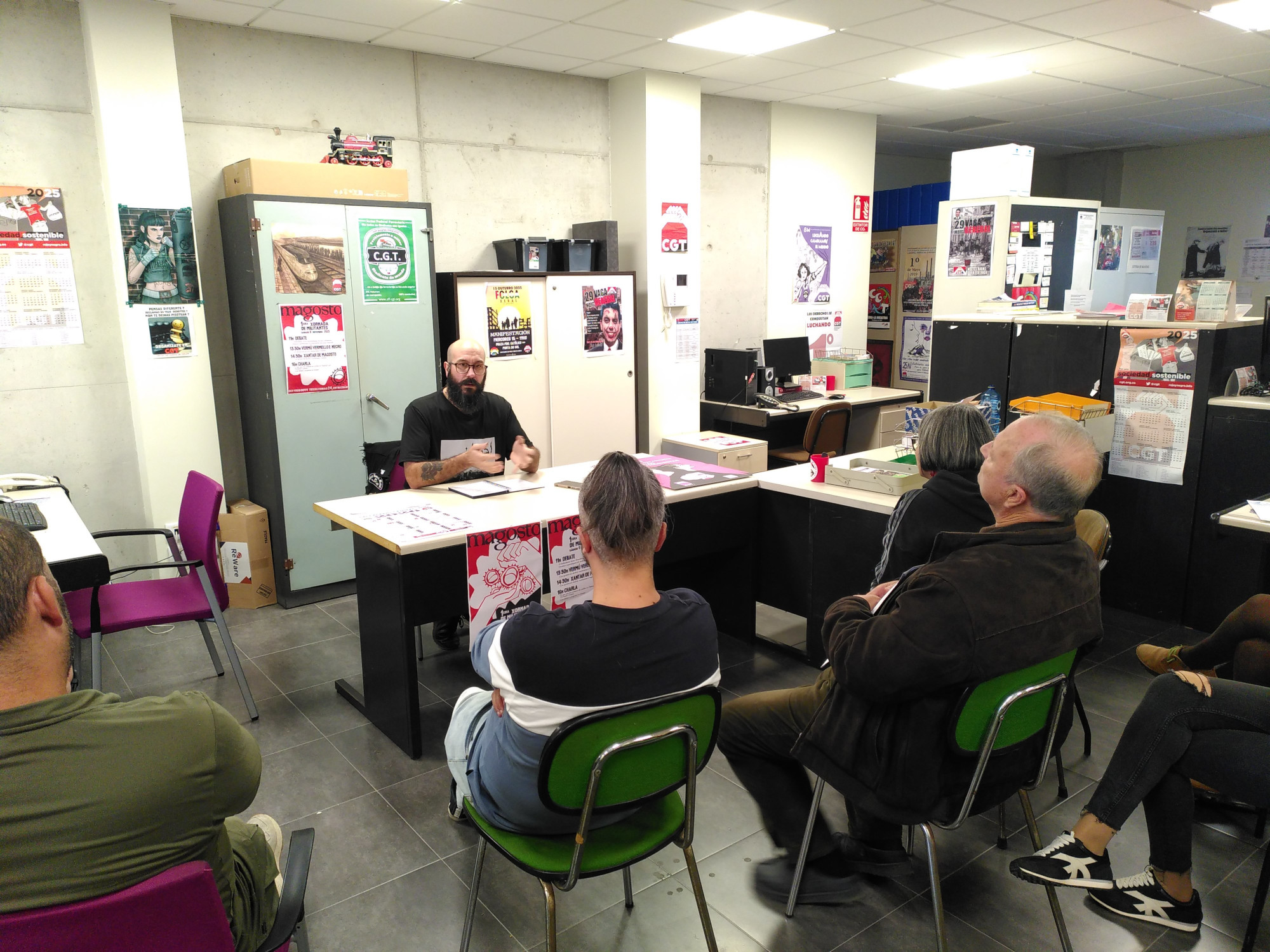 Na oficina de CGT Vigo, Fernando Carpizo imparte a súa charla "O que non che contaron do conflito en Palestina" sentado detrás dun escritorio. Varias persoas están sentadas diante del, escoitando atentamente.