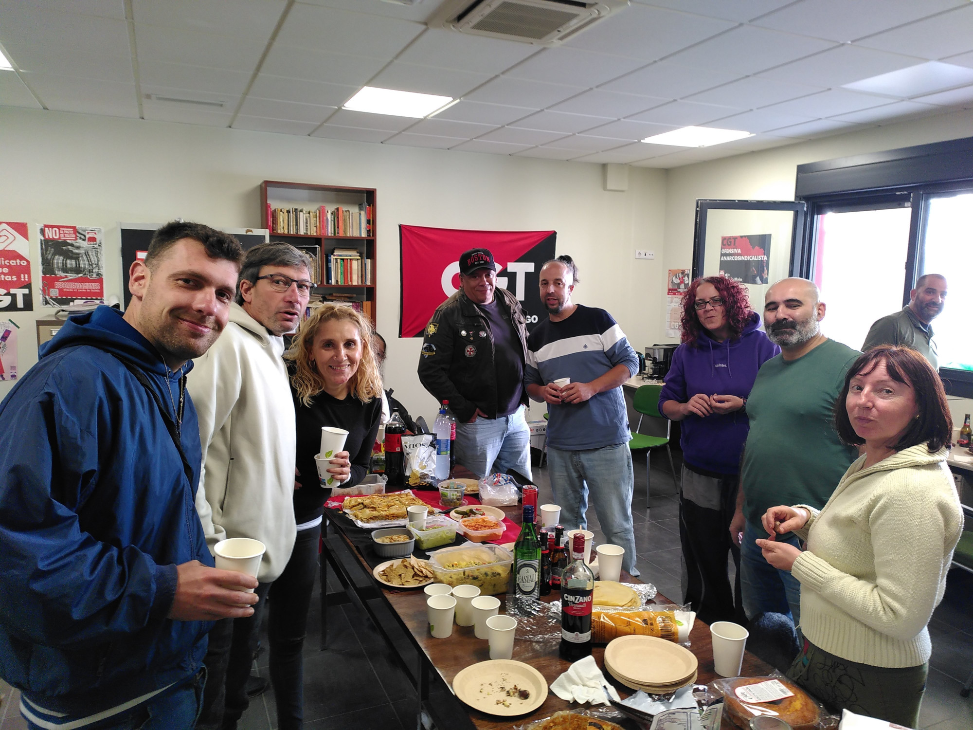 Un grupo de persoas de pé no local de CGT Vigo, mirando á cámara, sorrindo ou conversando nun ambiente relaxado e social. Están reunidas arredor dunha longa mesa de madeira chea de comida e bebida, incluíndo aperitivos, empanadas, salsas, botellas de refrescos, e vasos e pratos de papel. No fondo hai estanterías e carteis con texto e gráficos nas paredes, incluíndo unha bandeira vermella e negra co logotipo da CGT Vigo.
