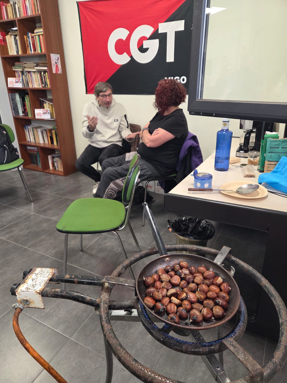 Interior do local da CGT Vigo. Nun primeiro plano, unha tixola asando castañas enriba dun queimador de gas portátil. No fondo, a secretaria de Acción Social charla cun afiliado diante da bandeira vermella e negra da CGT Vigo que está colgada na parede detrás deles.